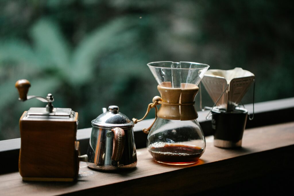 Manual coffee grinder with kettle served on wooden table with chemex and cup with paper filter on terrace overlooking green forest